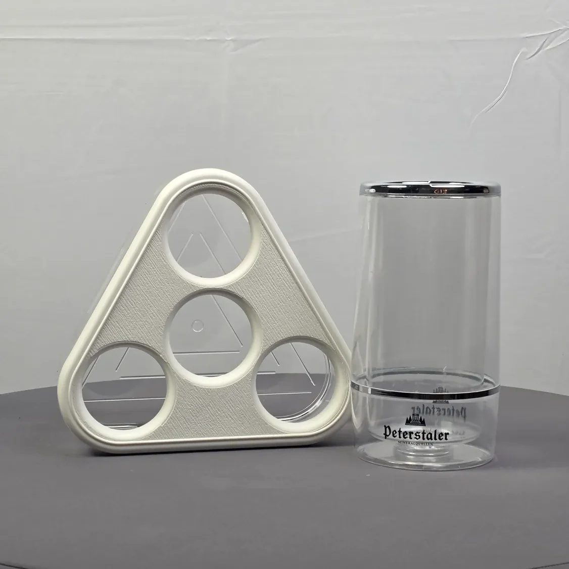 Vier runde Ausschnitte in einer weißen, dreieckigen Halterung neben einem durchsichtigen Behälter.