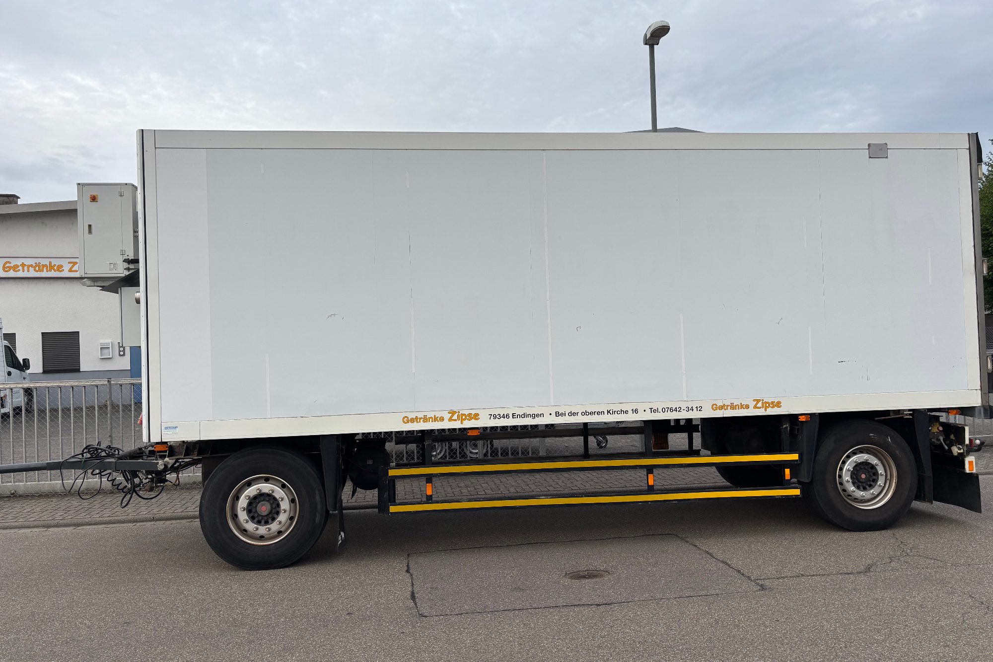 Weißer Lieferwagen mit Anhänger auf einer Straße, im Hintergrund Gebäude.
