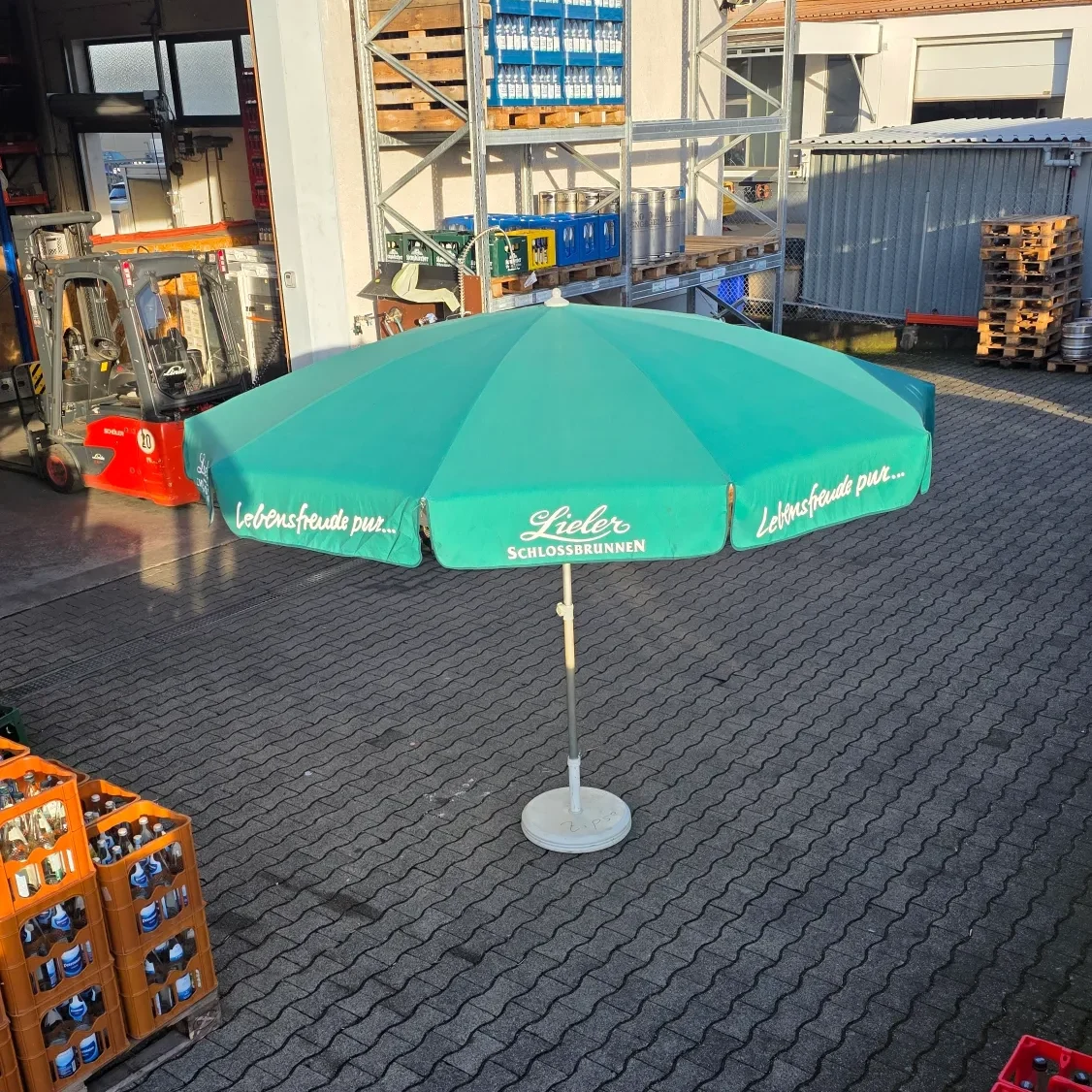 Schirm "Groß" Grüner Sonnenschirm auf einem gepflasterten Platz, umgeben von Kisten und Regalen.