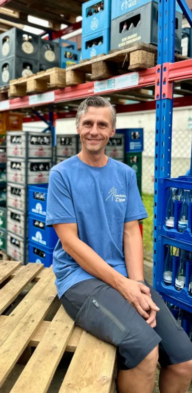 Mann in blauer T-Shirt sitzt auf Holzpalette, umgeben von Getränkekästen.