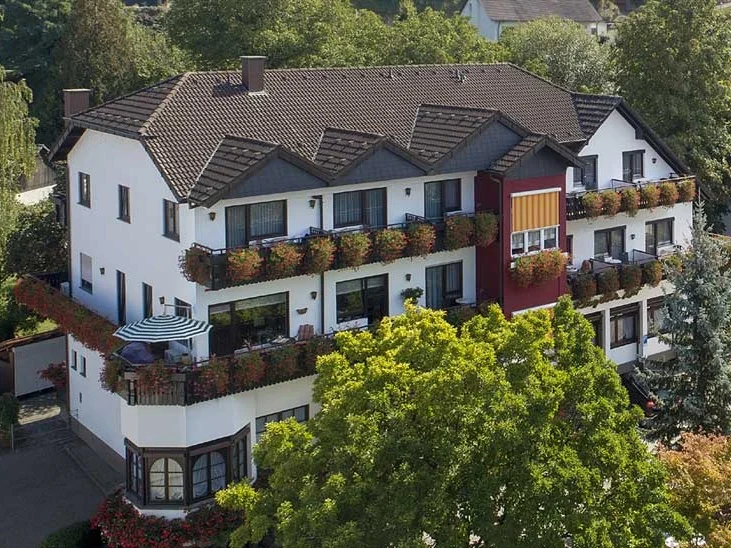 Ein mehrstöckiges, modernes Wohnhaus mit Balkon und vielen Blumen. Grüne Bäume umgeben es.