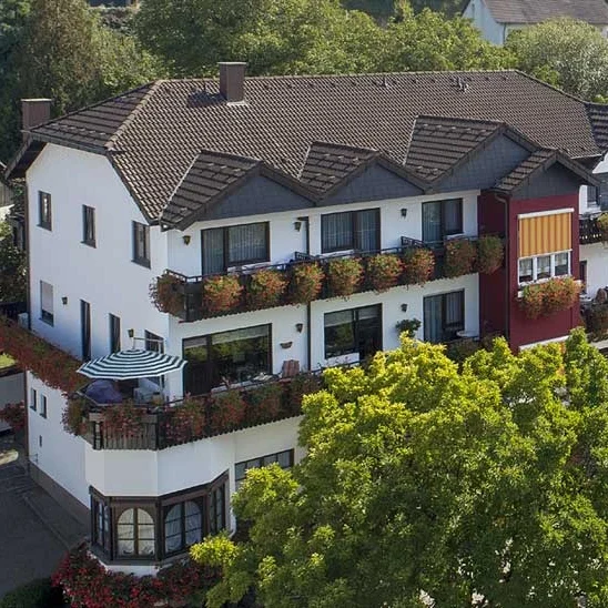 Mehrstöckiges Haus mit Balkon, umgeben von Bäumen und bunten Blumen.