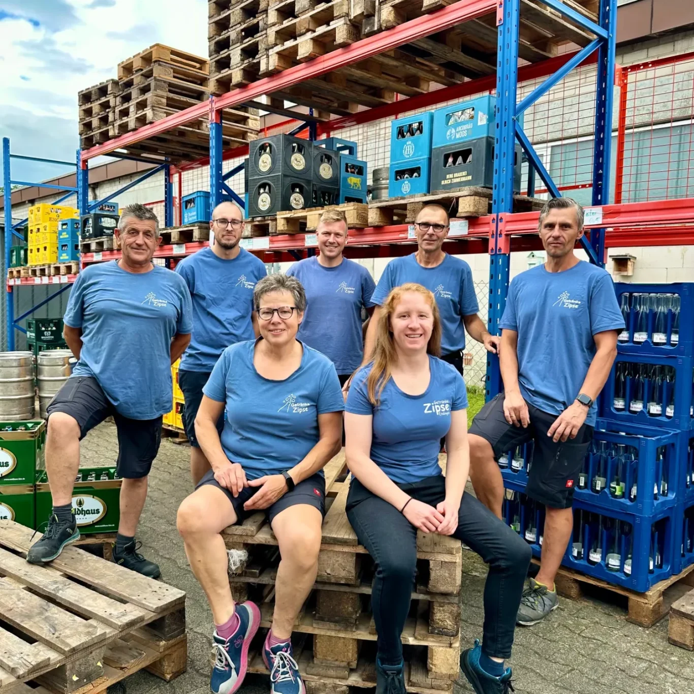 Sechs Personen in blauen T-Shirts sitzen und stehen zwischen Getränkekästen und Paletten.